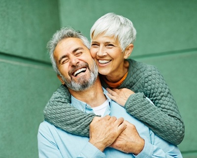 Smiling older woman hugs a laughing older man from behind. Happy couple shares a playful moment against a green wall.